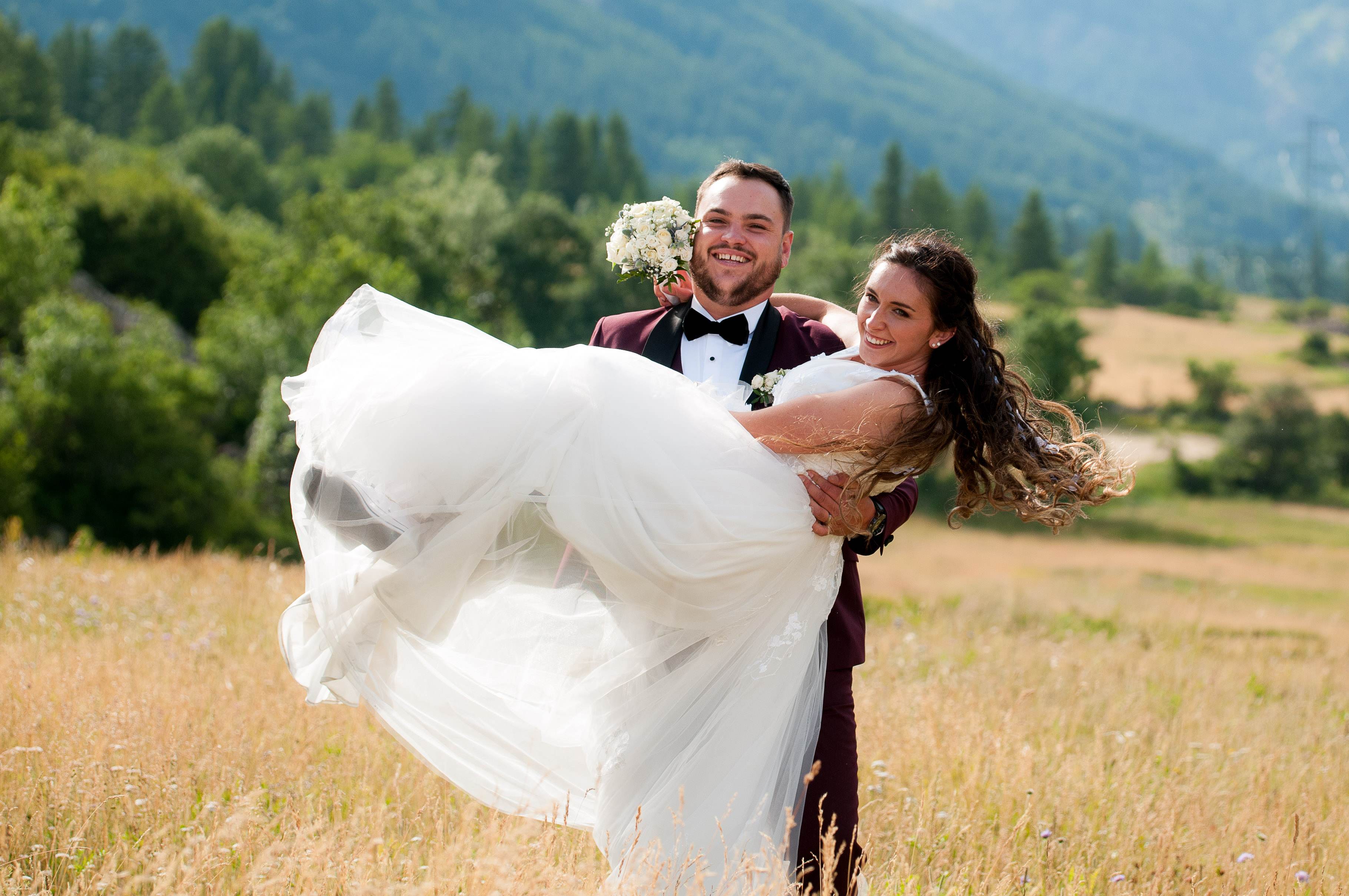 Le marié porte la mariée dans un pré, versants boisés des Alpes